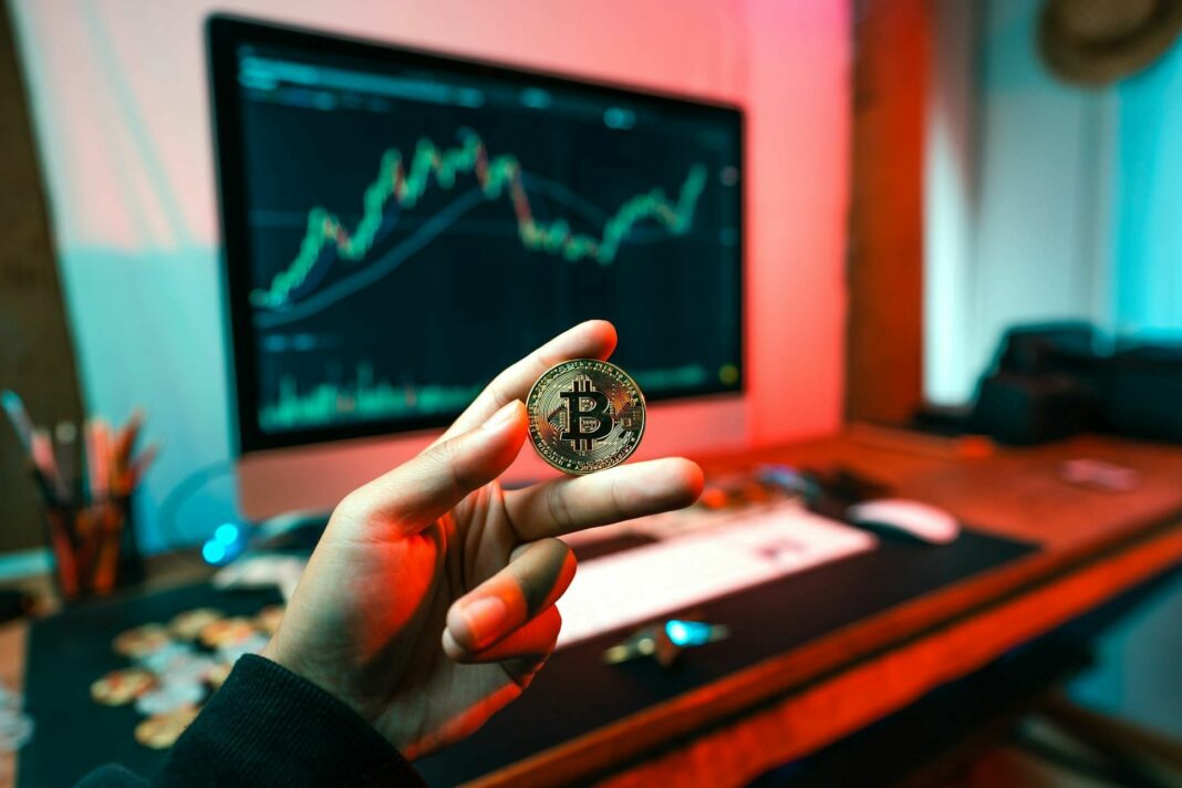 Main tenant une pièce Bitcoin avec écran d'analyse de marché crypto en arrière-plan